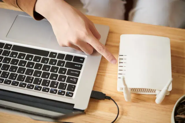 Dłoń wskazuje na router, pokazując, jak połączyć komputer z wifi. Kabel USB łączy laptop z urządzeniem.
